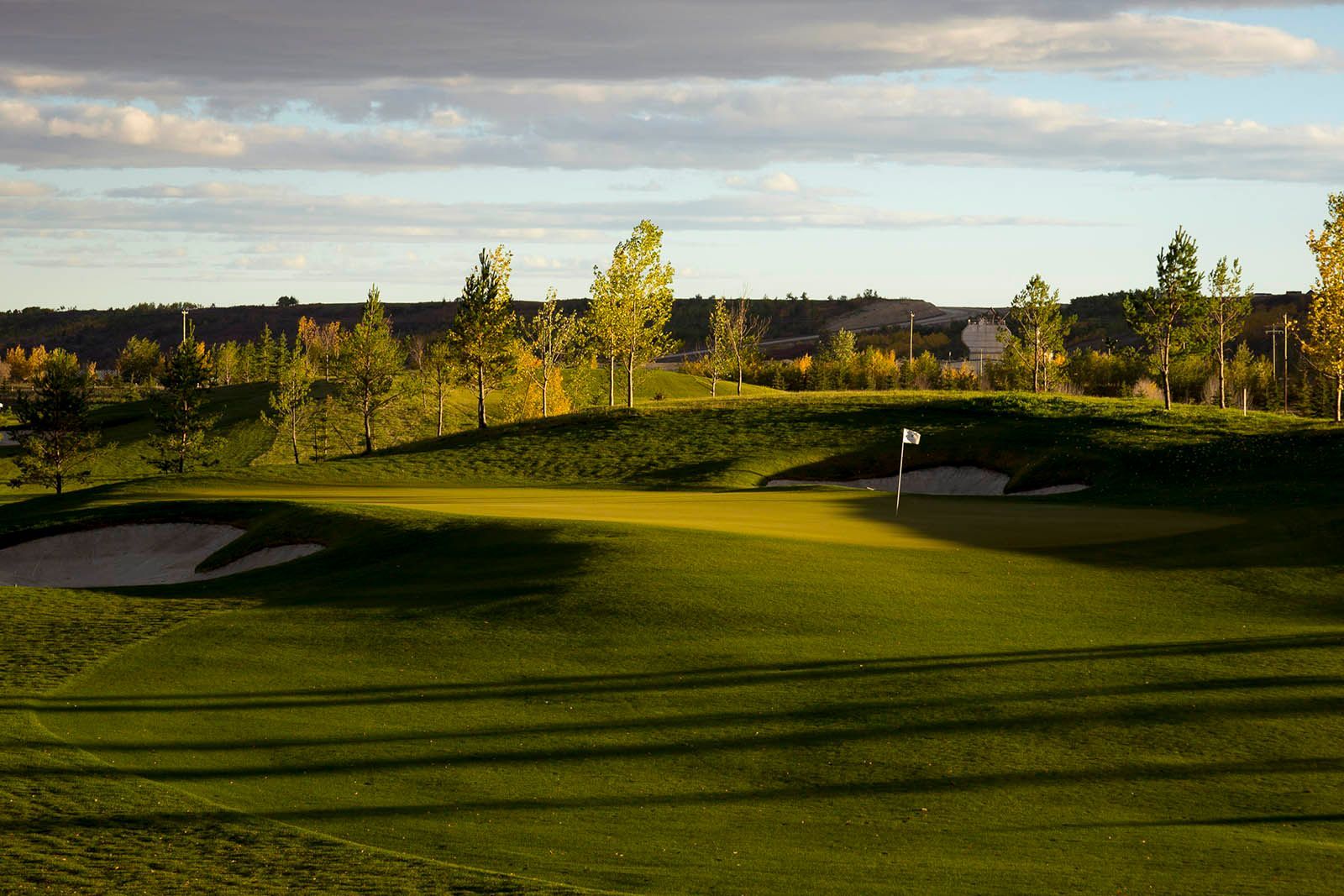 Green golf course with sand traps and flag, trees in the background under a cloudy sky.