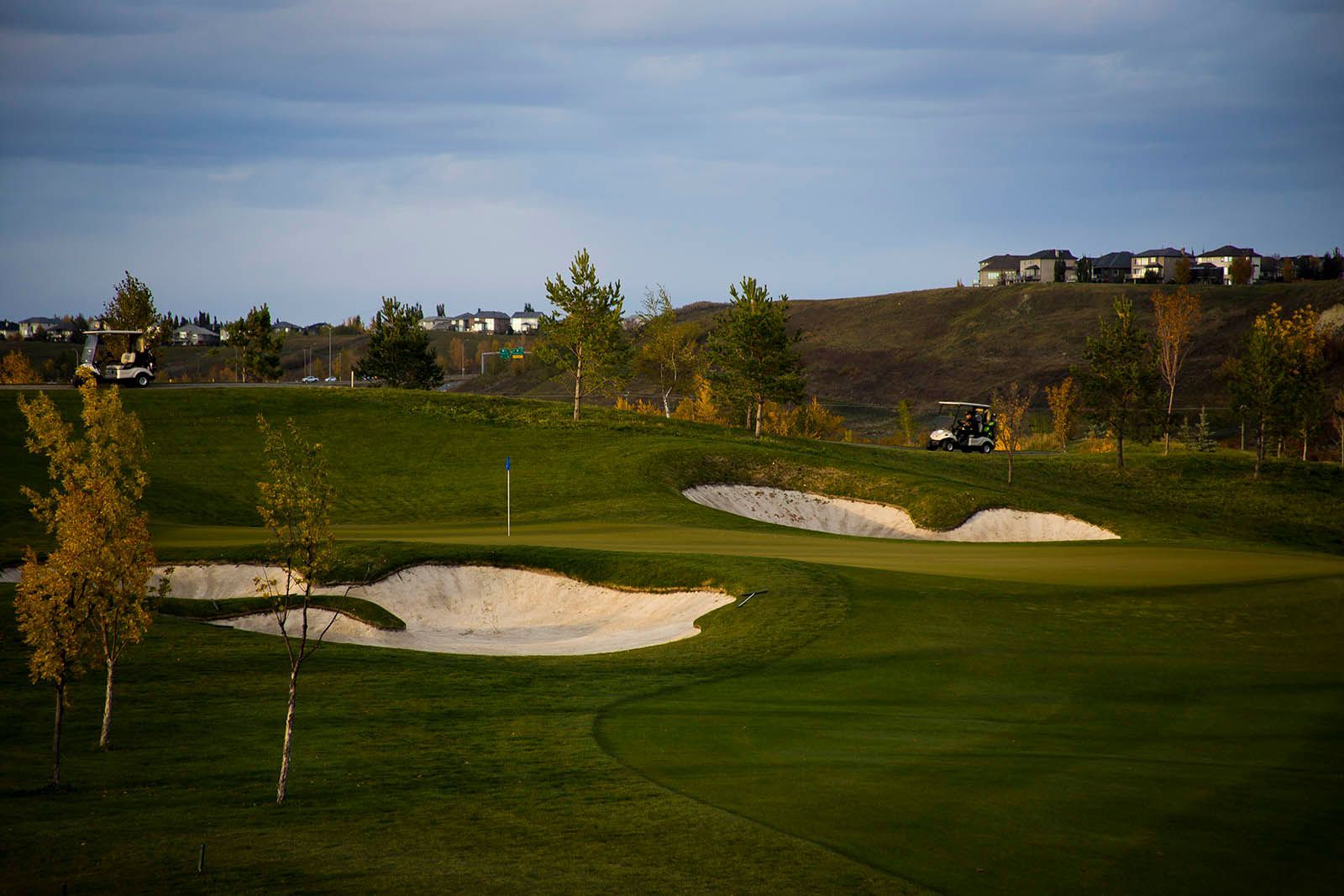 Golf course with bunkers, trees, and golf carts under a cloudy sky.