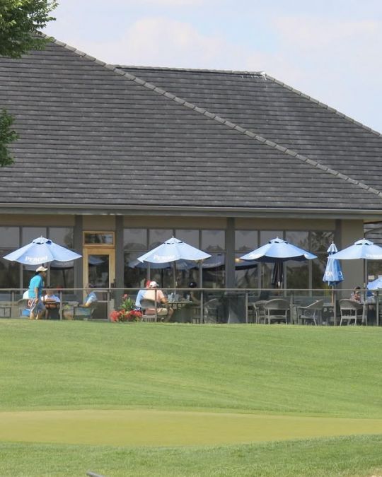 Building with shaded outdoor seating, overlooking a green golf course. People under blue umbrellas.
