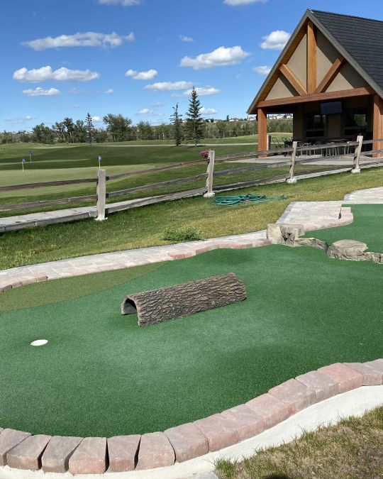 Mini golf course with green turf, a log tunnel, and a wooden gazebo under a blue sky.