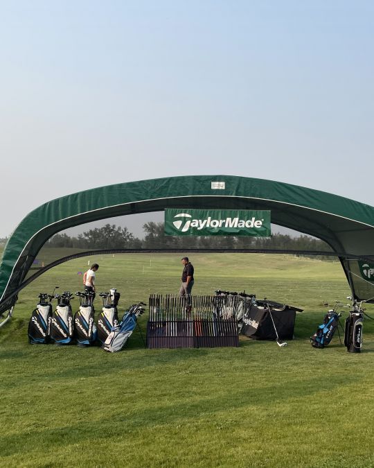 Golf club display under a TaylorMade tent on a green field with two people.