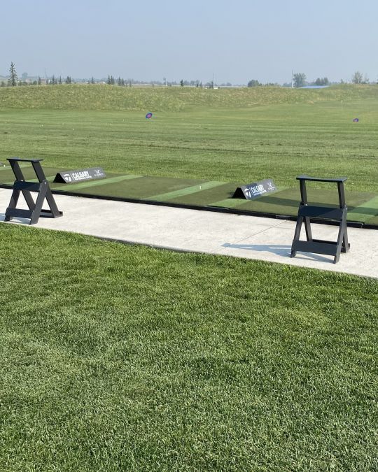 Golf driving range with two black chairs on a concrete pad, green grass and field in the background.