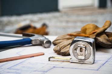 A tape measure , gloves , a hammer , and a pencil are on a blueprint.