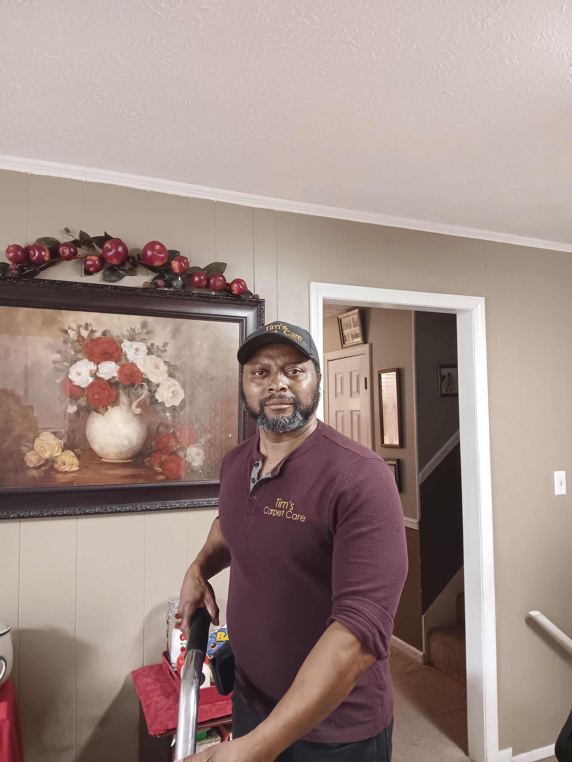 Man in burgundy shirt and cap, holding an object, standing near a framed painting.