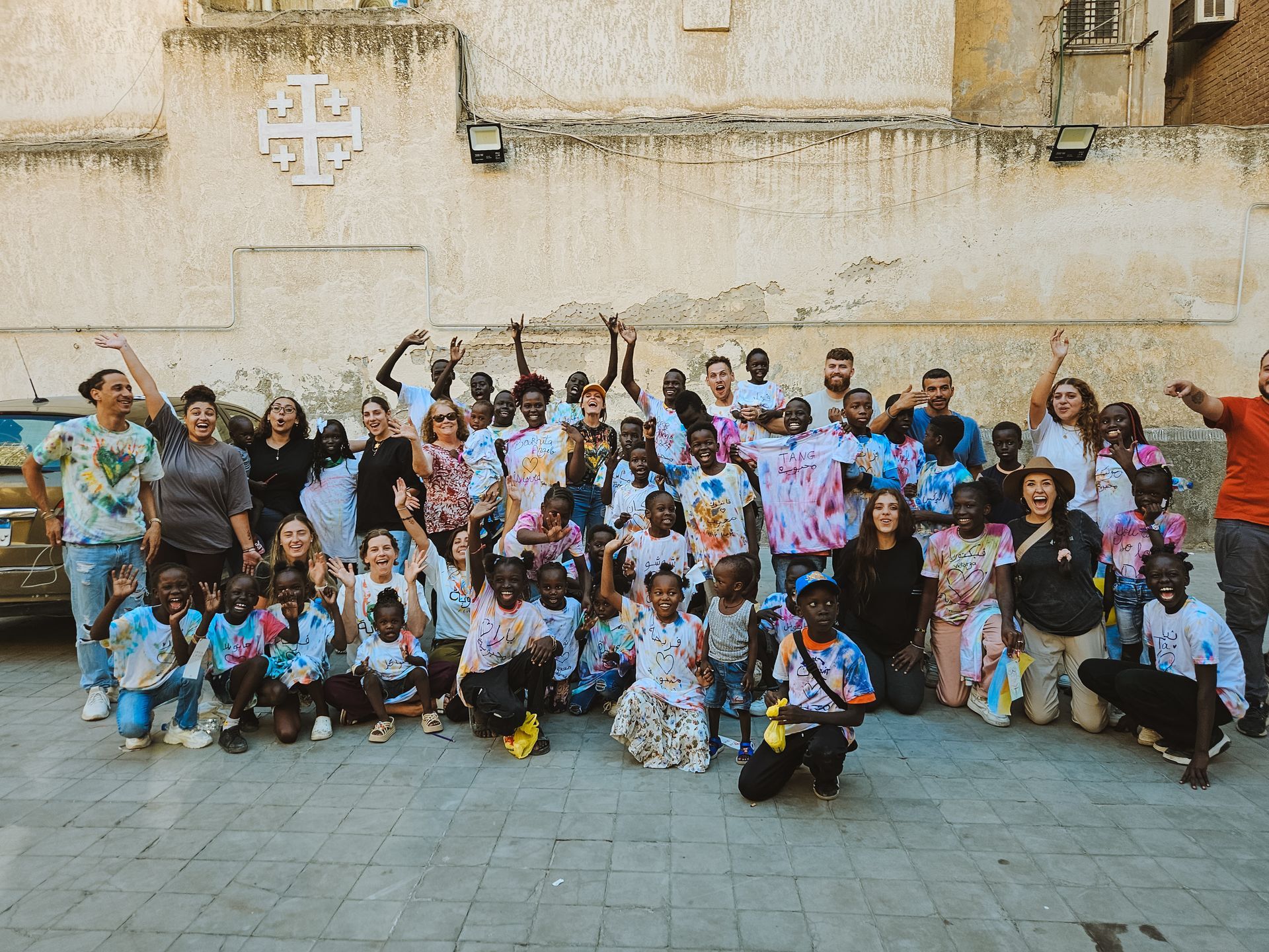 Group of people, mostly children, posing together outdoors; some are raising arms. A building is in the background.