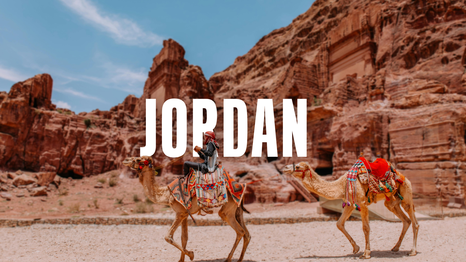 Two camels with riders travel across a sandy path in front of the red sandstone cliffs of Petra, Jordan.