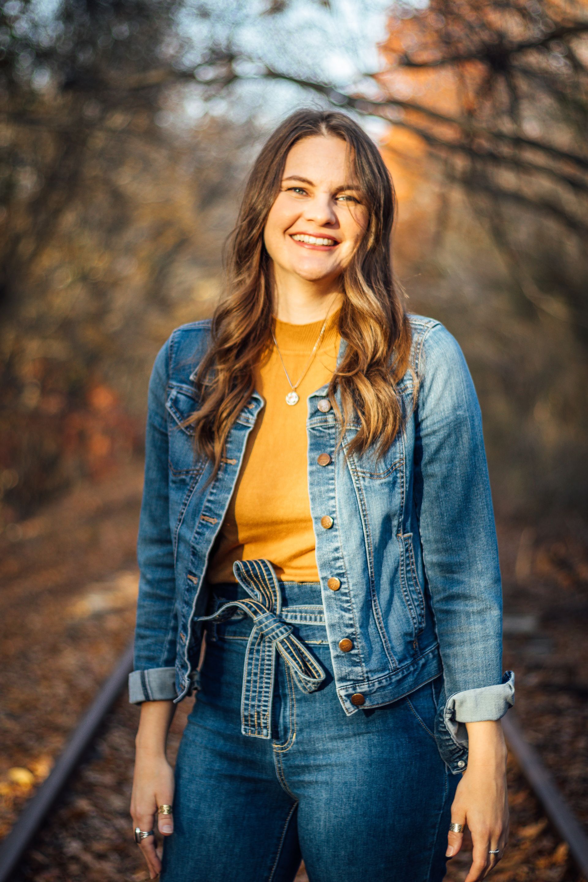 Woman in denim jacket and jeans smiles on railroad tracks. Brown and orange fall foliage in background.