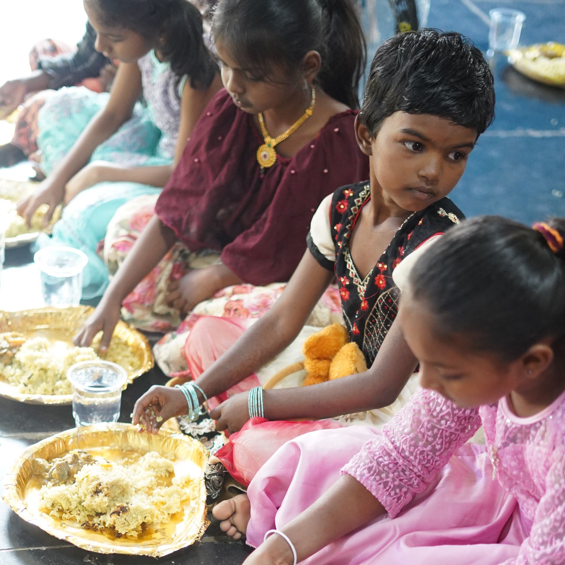 Children seated on floor, eating from plates. Food visible. Indoor setting.