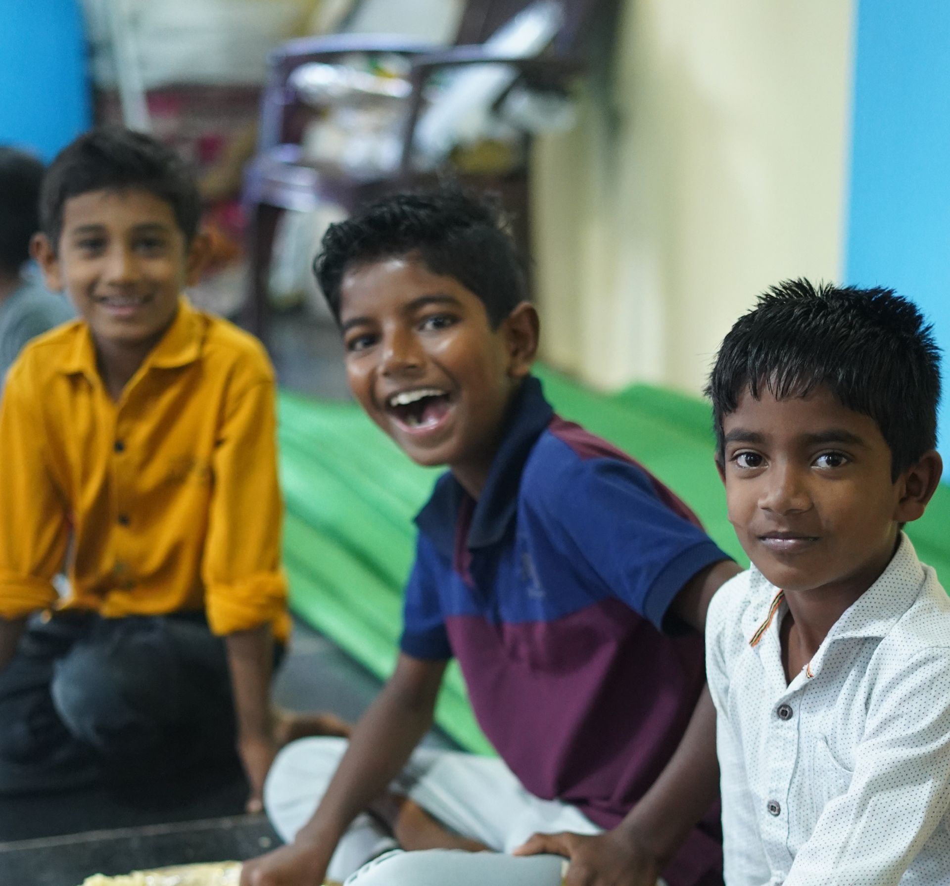 Three smiling children sit on the floor; one in yellow, one in blue, and one in white.