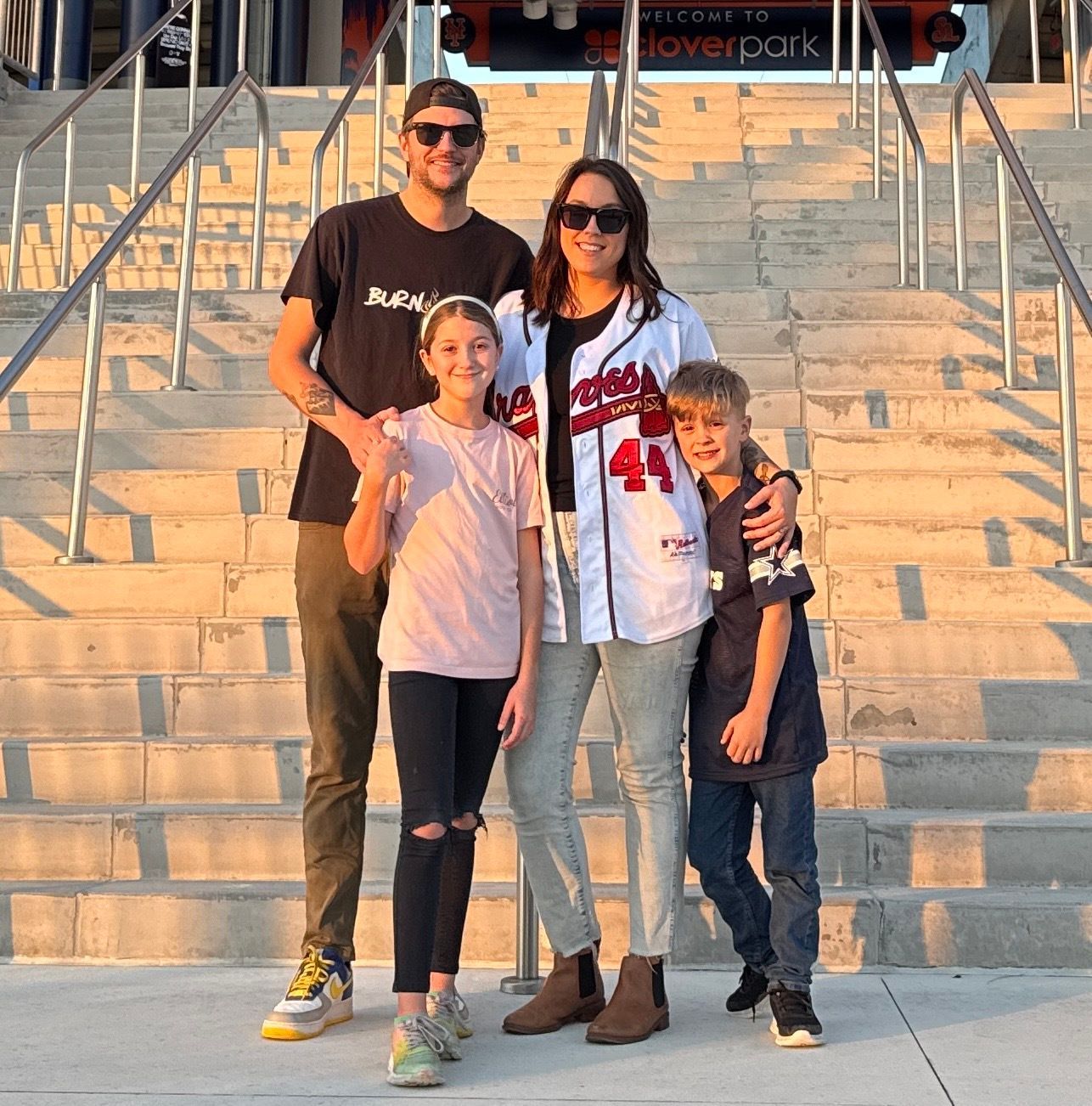 Family of four standing on stairs, smiling. The woman wears a baseball jersey, the man a black tee.