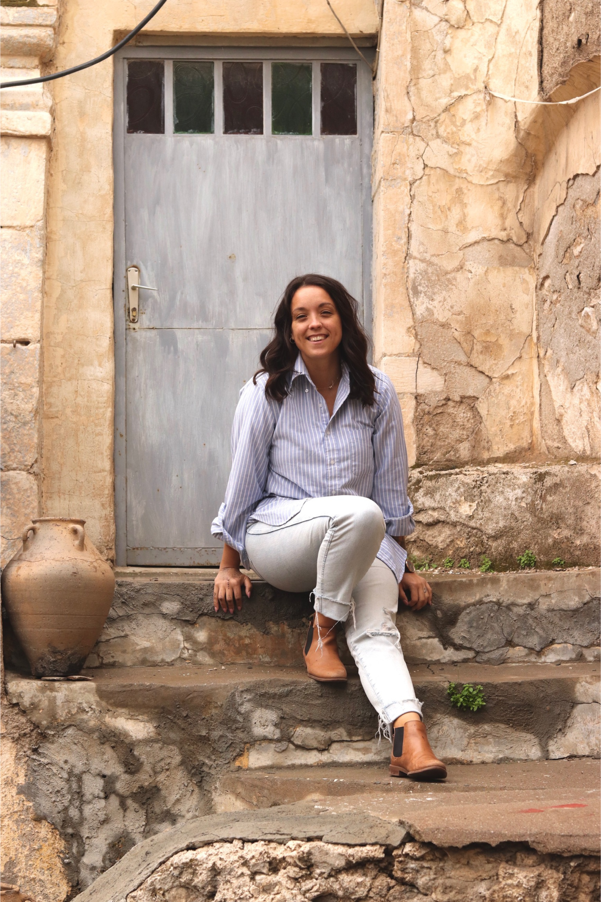 Woman sitting on stone steps in front of a blue door, wearing a striped shirt and white pants, smiling.