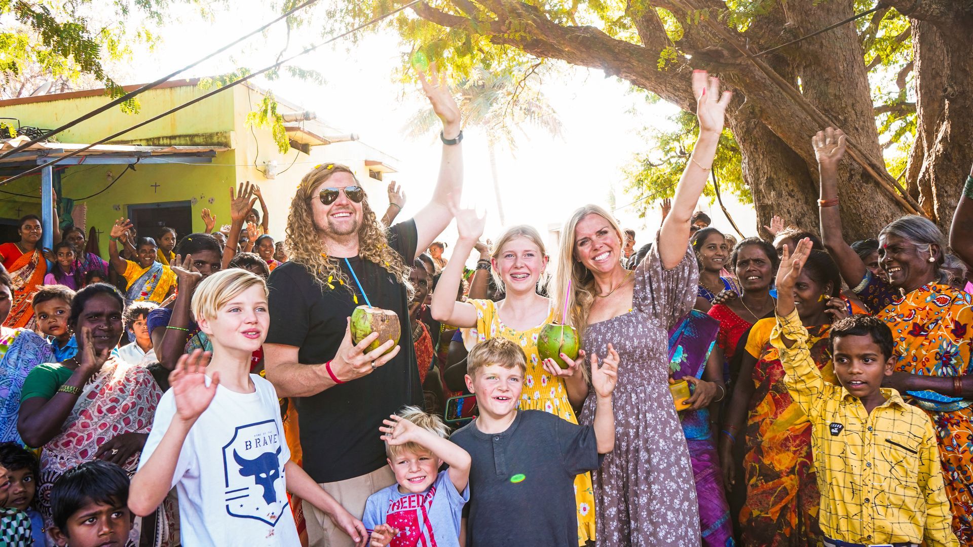 Family and village crowd waving, smiling, holding coconuts, under a tree.