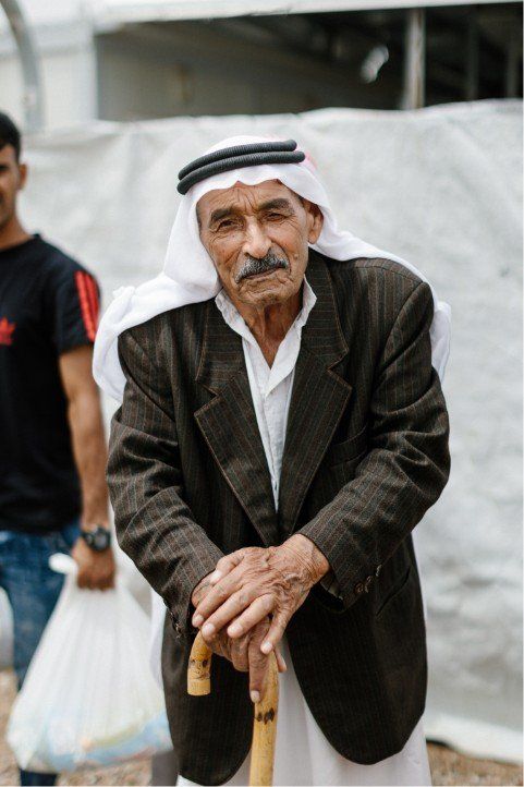 Older man in jacket and headscarf, holding cane, posing. Another person in background.
