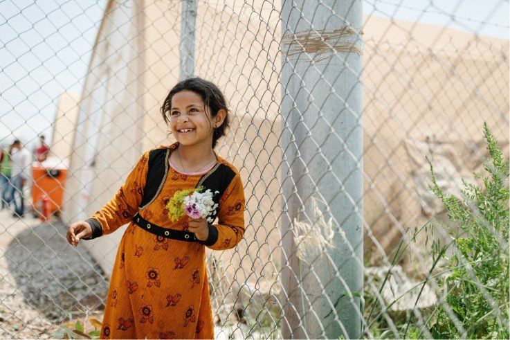 Girl smiles, holding flowers by a fence in front of a tent; sunlight.