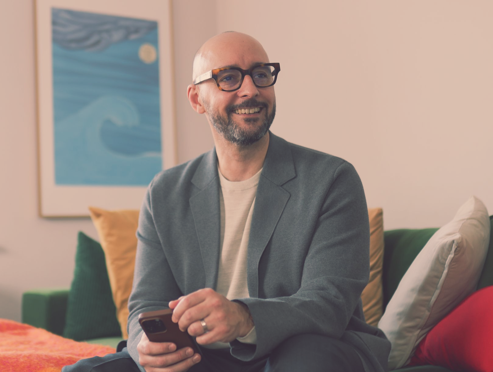 Justin Tate of The Exec Memo with glasses, gray blazer, and holding a phone, smiles while sitting on a couch.