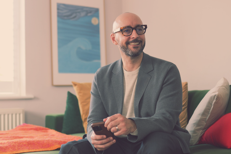 Justin Tate of The Exec Memo  with glasses, holding a phone, smiles while seated on a green couch in a living room.