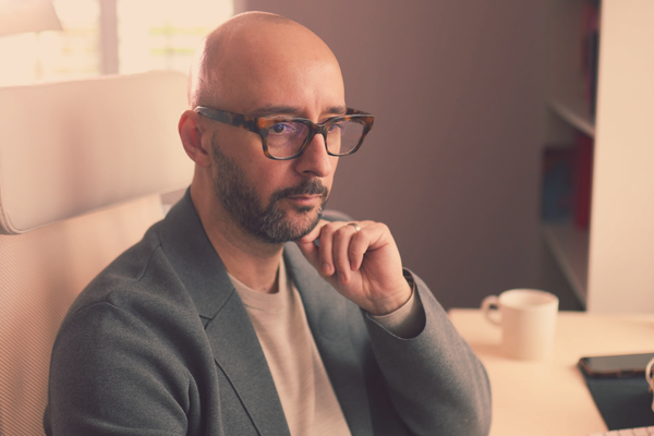 Justin Tate wearing glasses, resting chin, seated at desk, looking thoughtful.