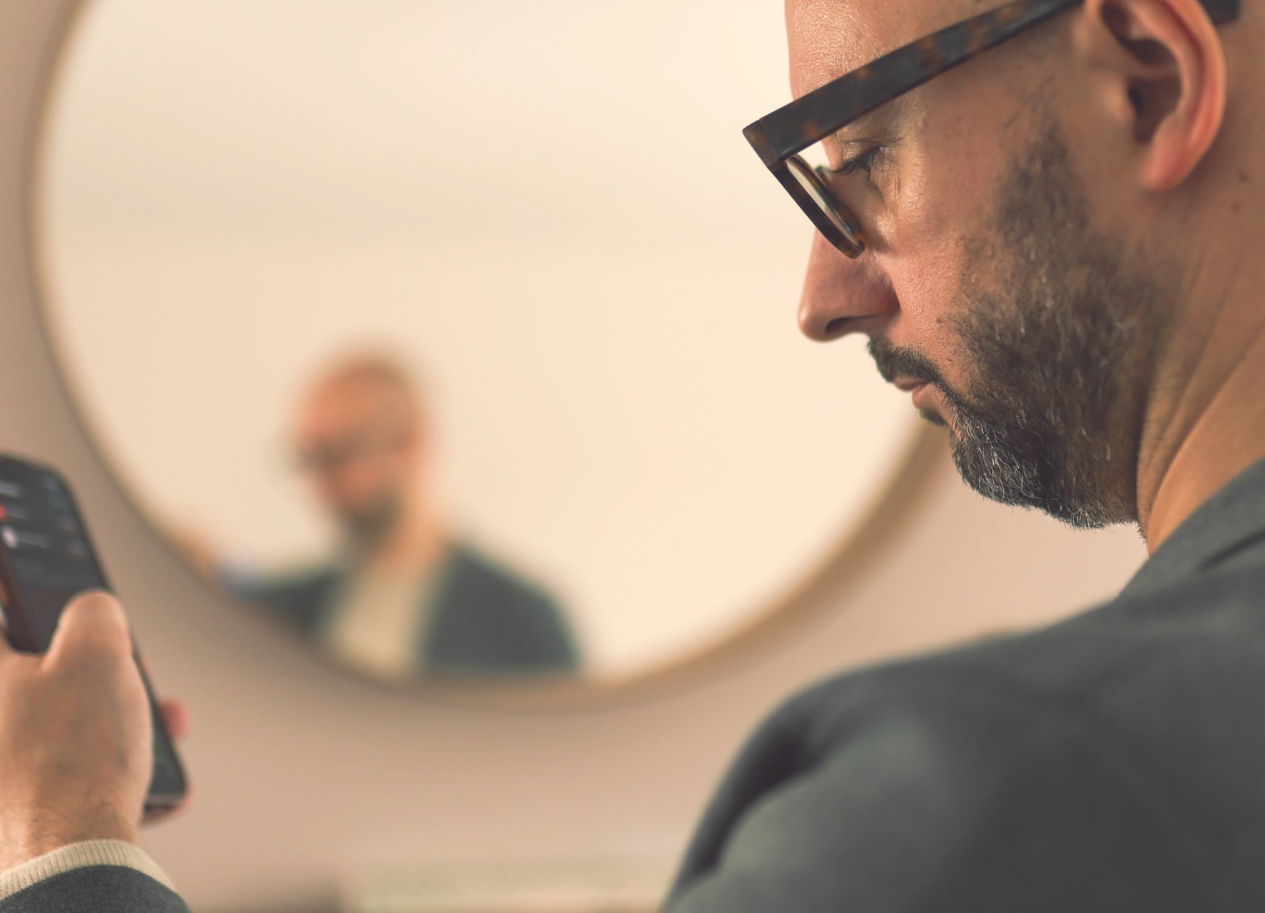 Justin Tate of The Exec Memo in glasses looking at phone, reflection in mirror shows blurred image of him.