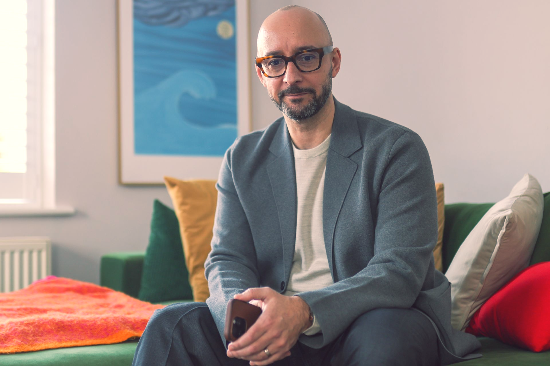 Justin Tate of The Exec Memo  with glasses, holding a phone, smiles while seated on a green couch in a living room.