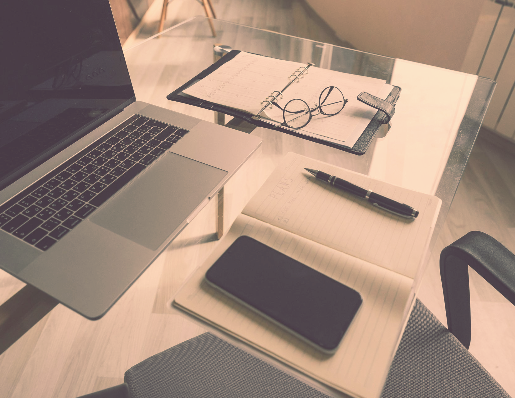 Laptop, notepad, pen, glasses, and phone on a glass desk, ready for work.