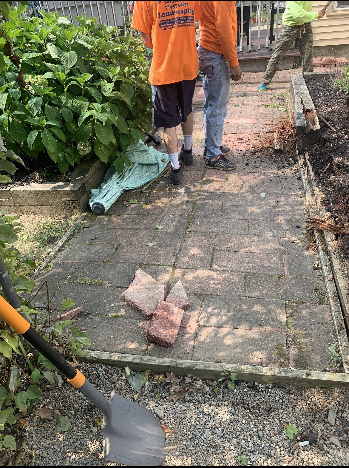 Two men are standing on a brick walkway next to a shovel.