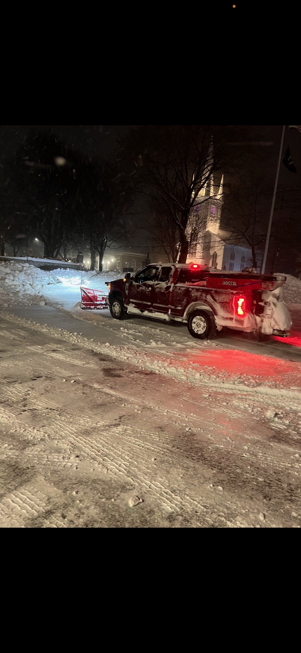 A red truck is driving down a snowy street at night.