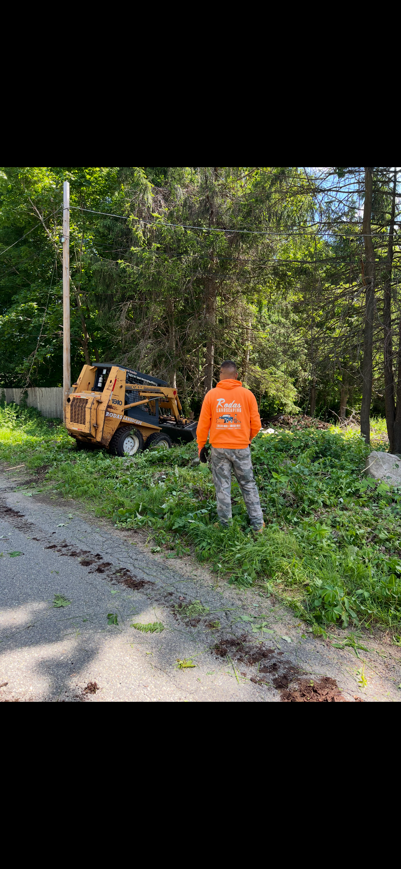 A man in an orange shirt is standing next to a tree stump grinder.