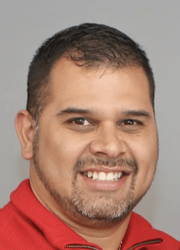 A man with a beard is wearing a red shirt and smiling. Ezzy Rodas