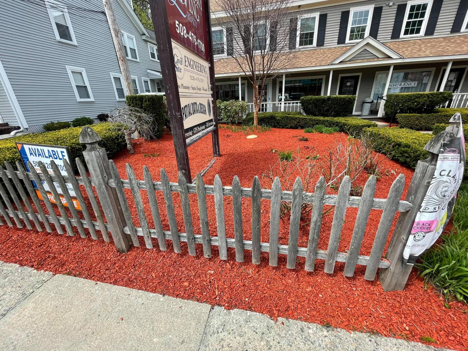 A wooden fence is surrounded by red mulch in front of a house.
