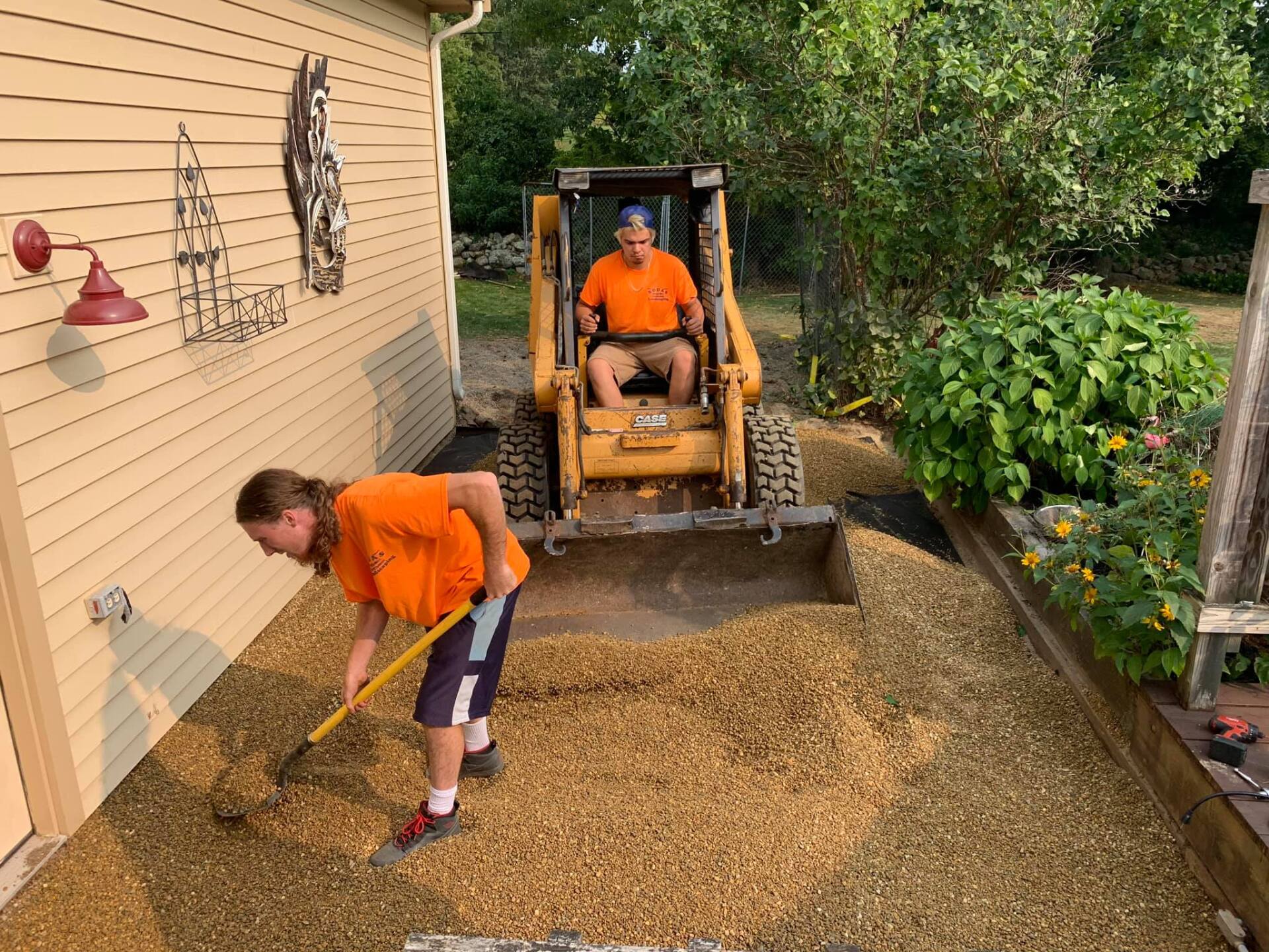 A man is driving a bulldozer and a woman is digging in front of a house.