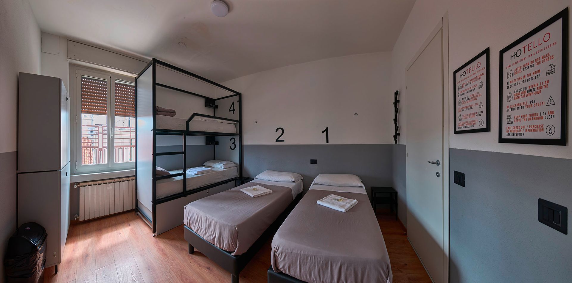 A hostel room with bunk beds, two twin beds, a window, and wall art. Grey and white color scheme.
