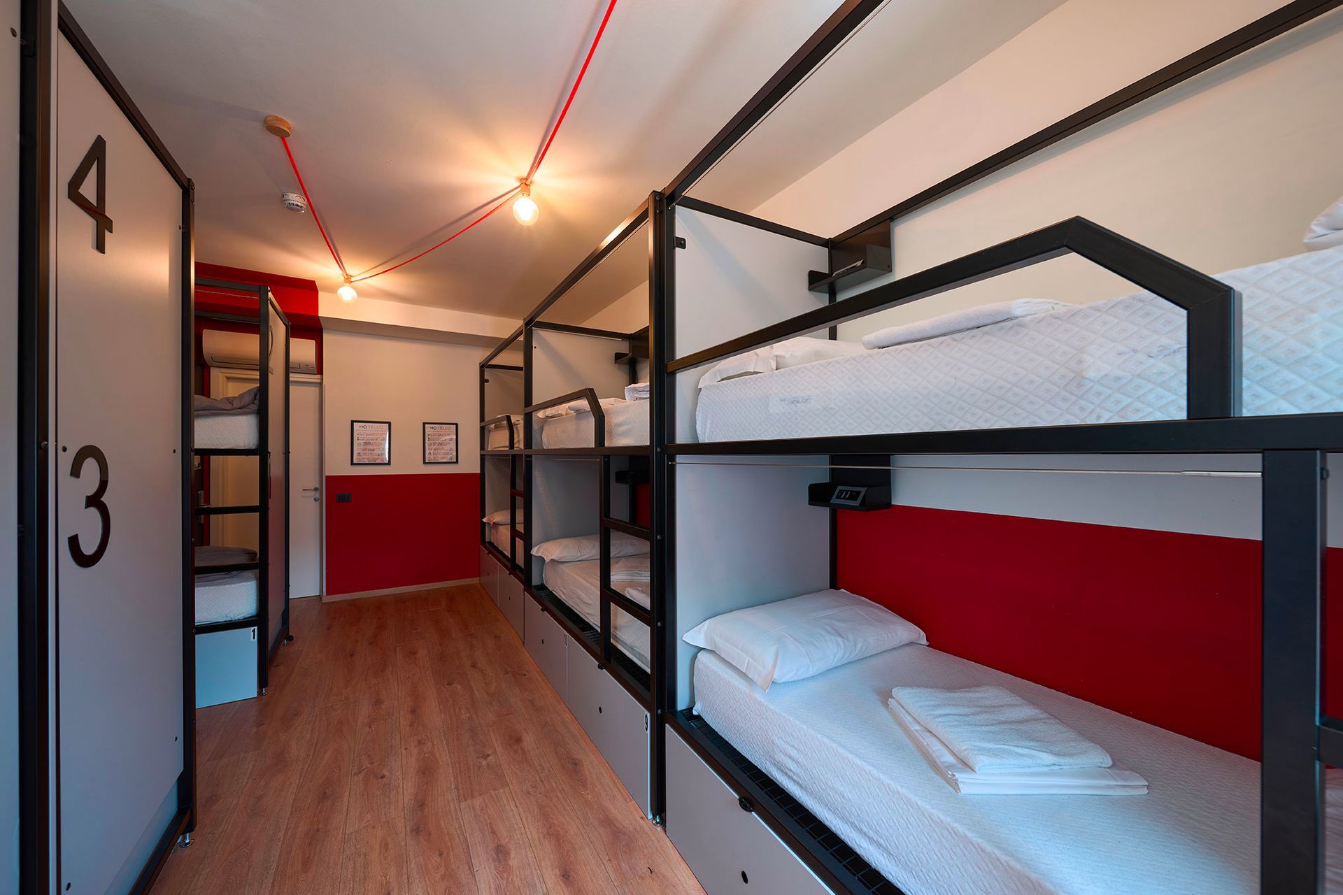 Hostel room with several bunk beds along a hallway; red accents, white linens.