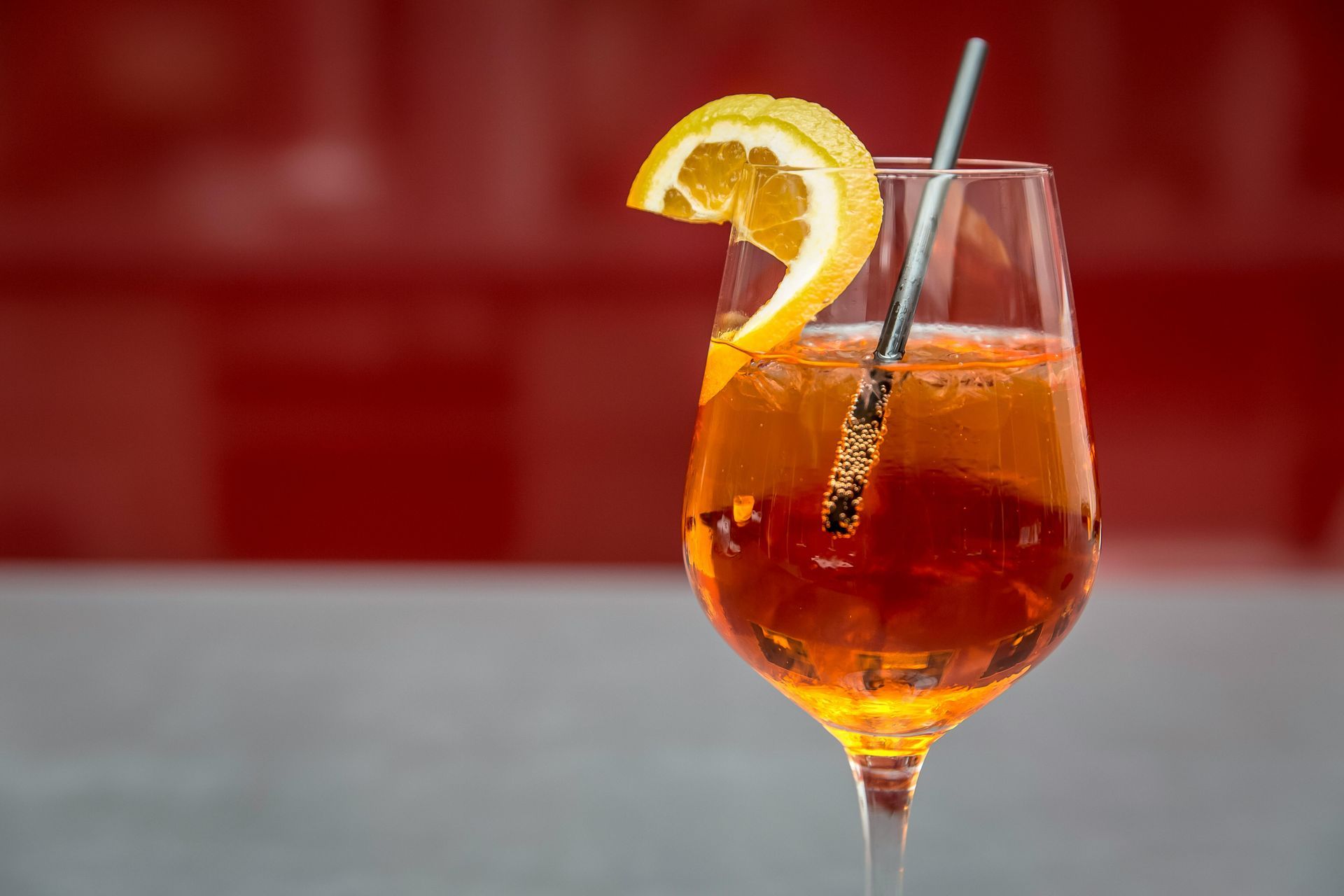 Spritz Aperol in un calice da vino, guarnito con una fetta d'arancia e una cannuccia di metallo, su uno sfondo rosso.