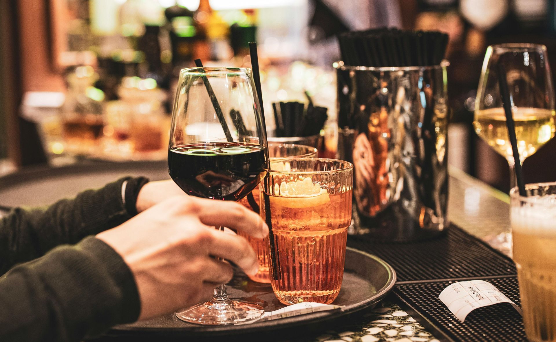 Persona che regge un vassoio con delle bevande, tra cui un bicchiere di vino rosso, in un ambiente da bar.
