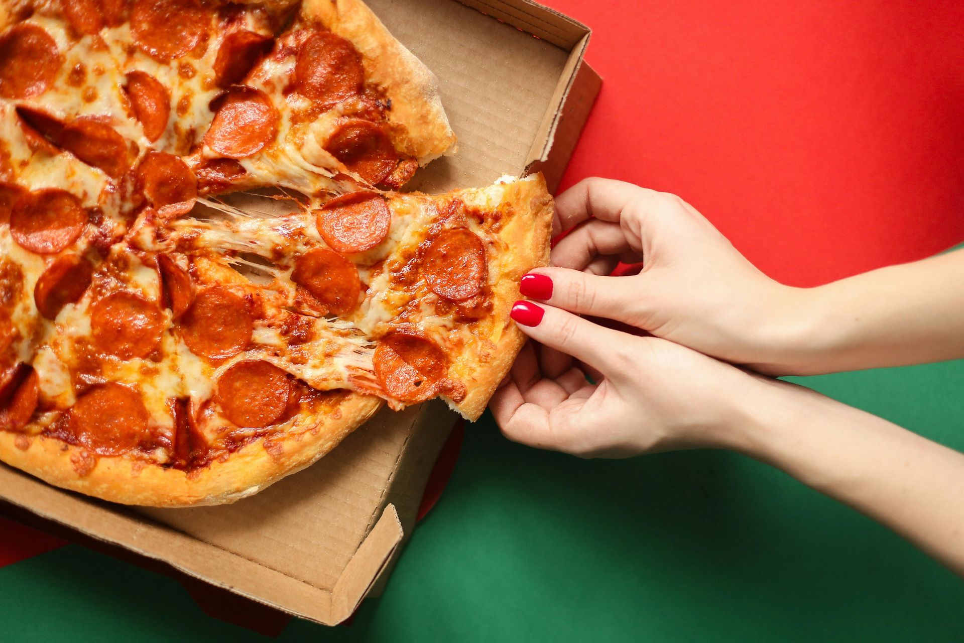 Persona che tiene in mano una fetta di pizza al salame piccante da una scatola, su sfondo rosso e verde.
