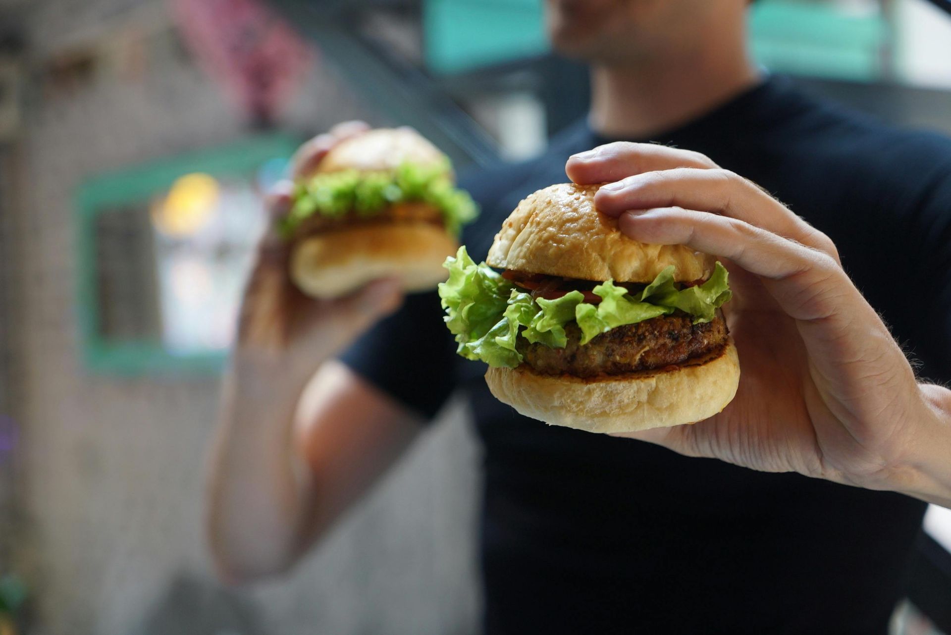 Persona che tiene in mano due hamburger con lattuga davanti a uno sfondo sfocato.