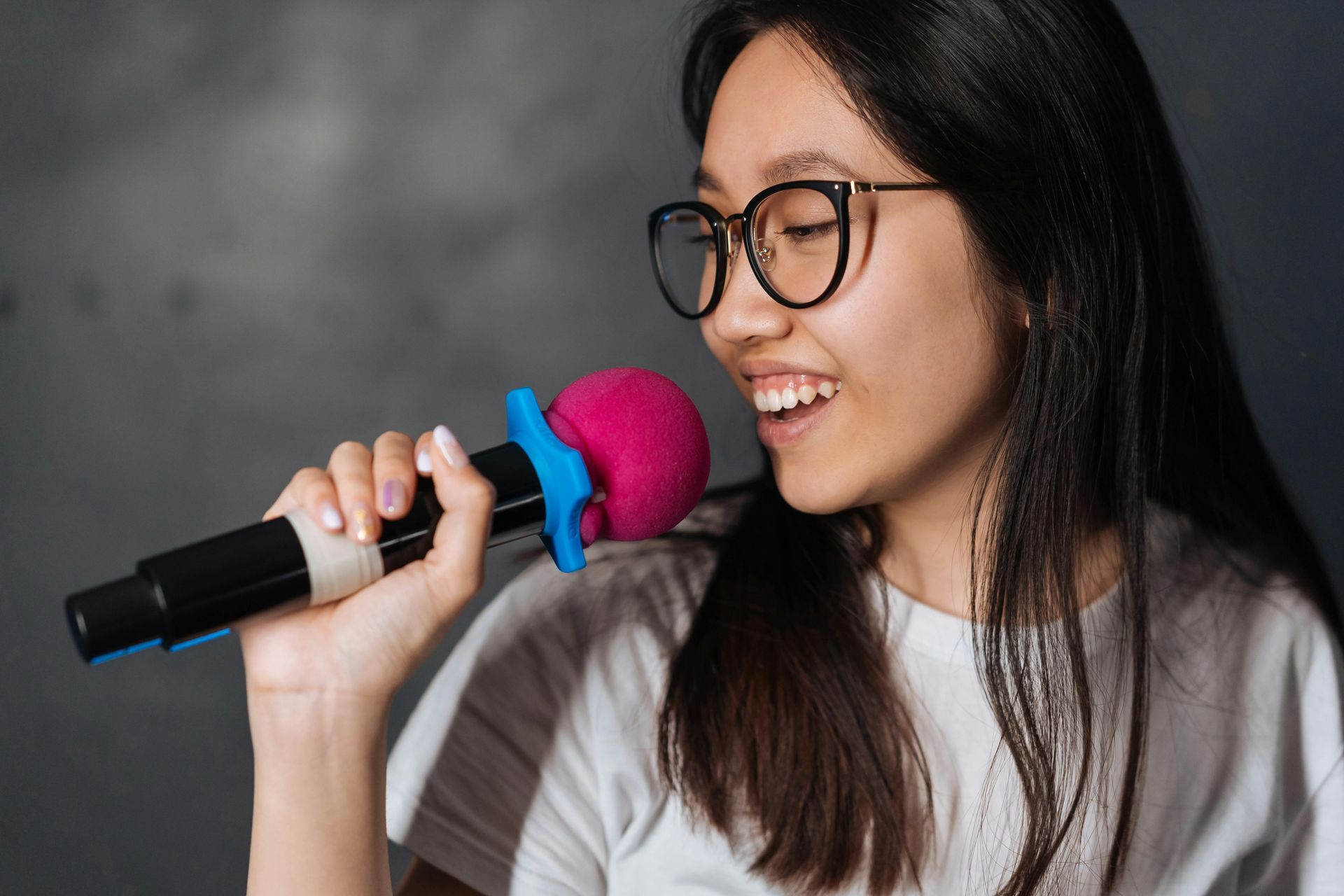 Giovane donna con gli occhiali che canta in un microfono; testa rosa, sfondo grigio.