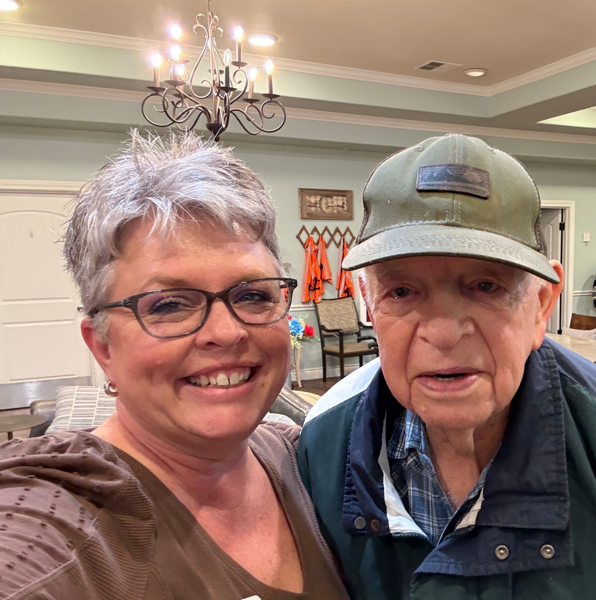 A smiling woman and a man in a green baseball cap pose together for a photo in a brightly lit indoor space.