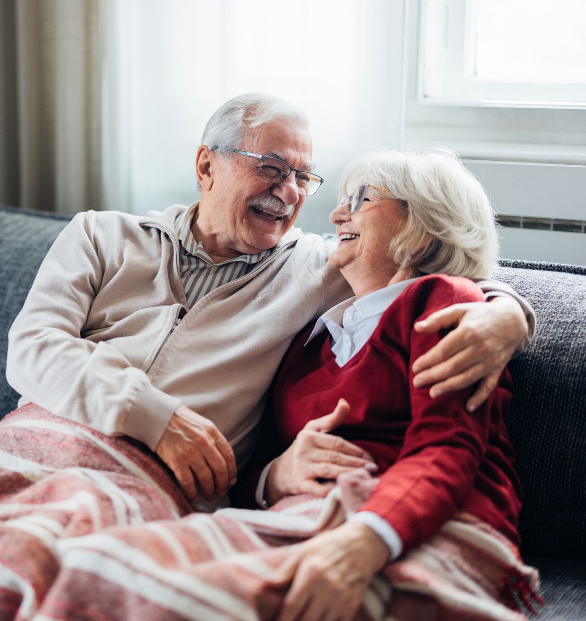 Two people with grey hair wearing glasses sit on a sofa, laughing together under a patterned blanket.