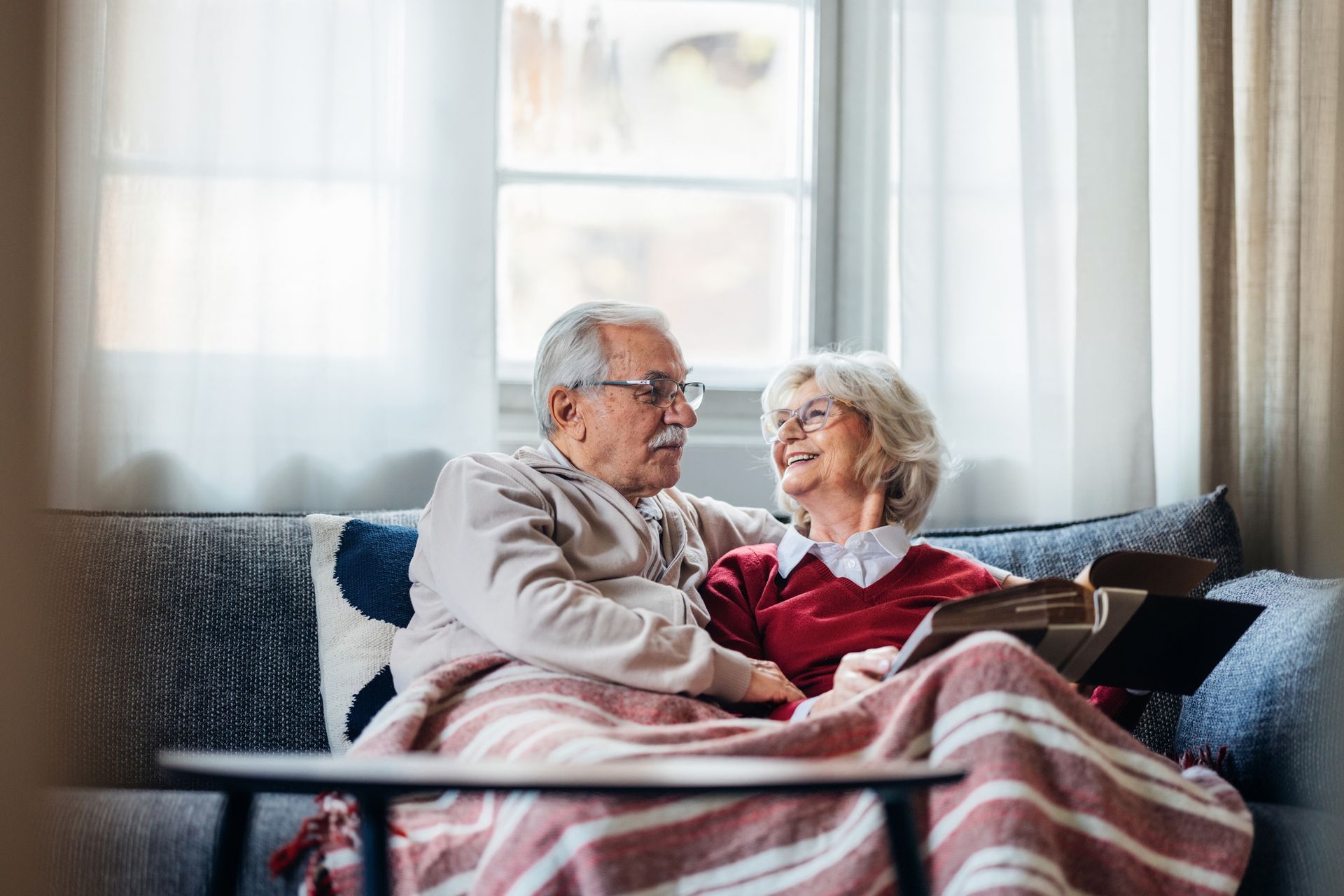 Two people sit together on a sofa under a striped blanket, smiling at each other while looking at a book.