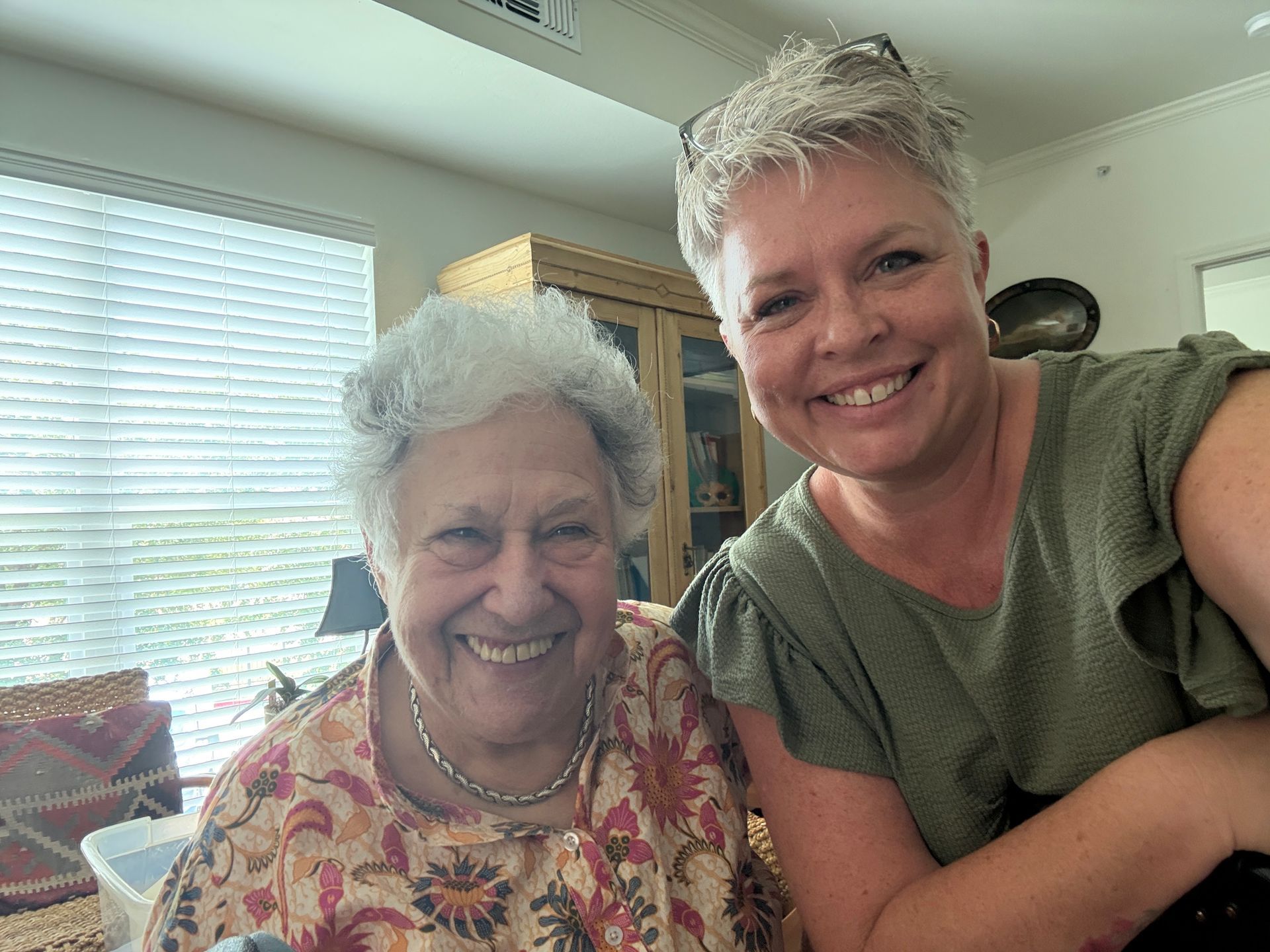 Two people smiling for a selfie in a sunlit room, one wearing a patterned floral top and the other an olive green shirt.