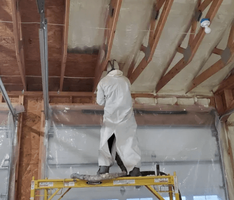 Person in protective suit spraying foam insulation on a wall inside a building.