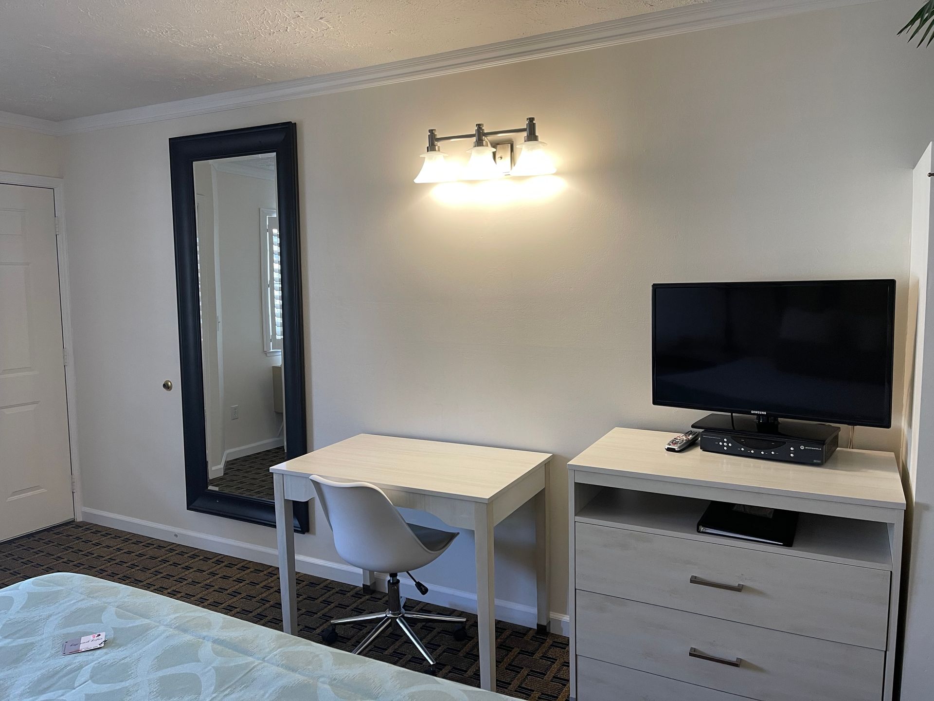 Bedroom with a desk, dresser, mirror, and TV against a white wall.