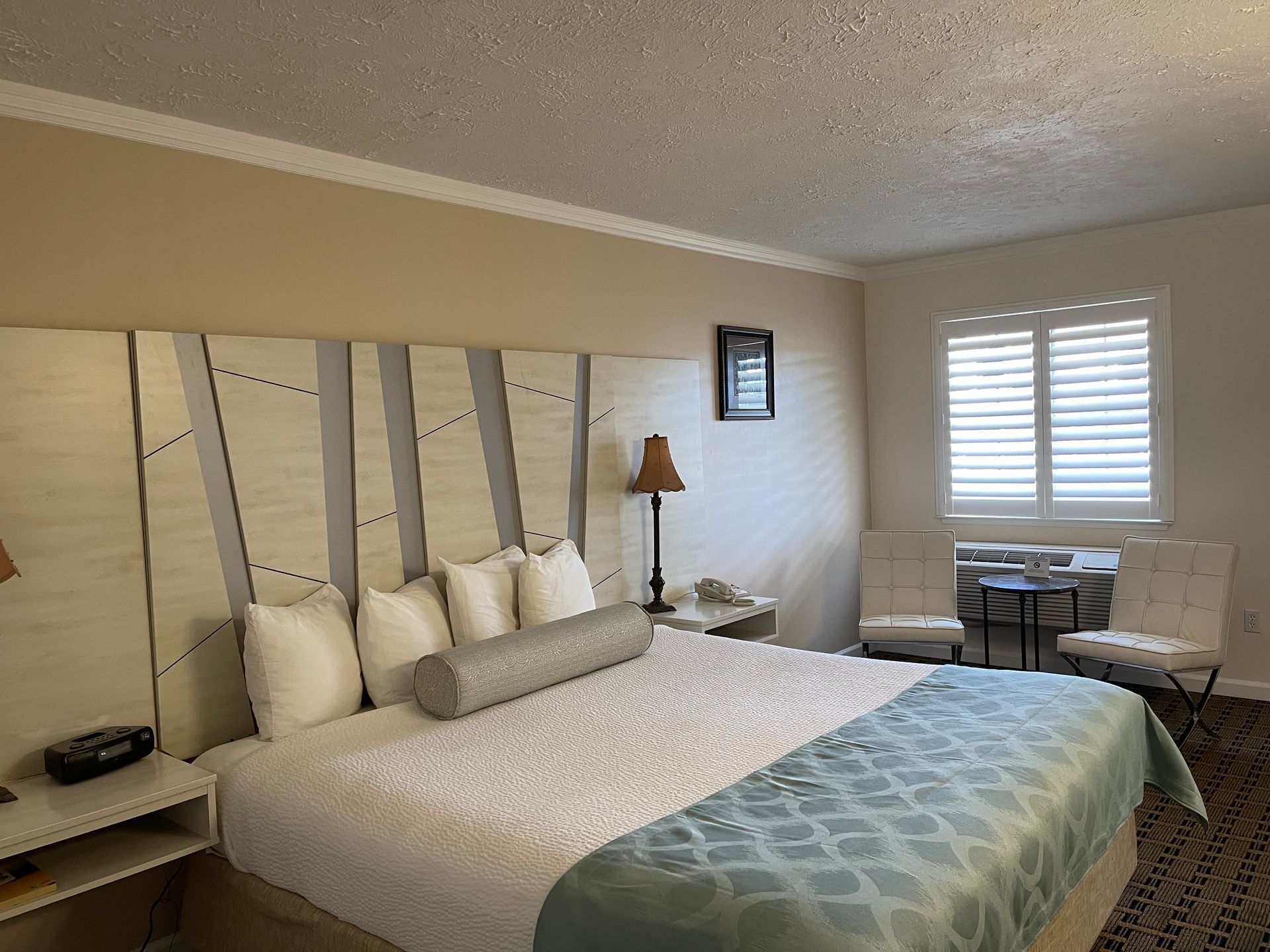 Hotel room with king-size bed, patterned headboard, bedside tables, and chairs near a shuttered window.