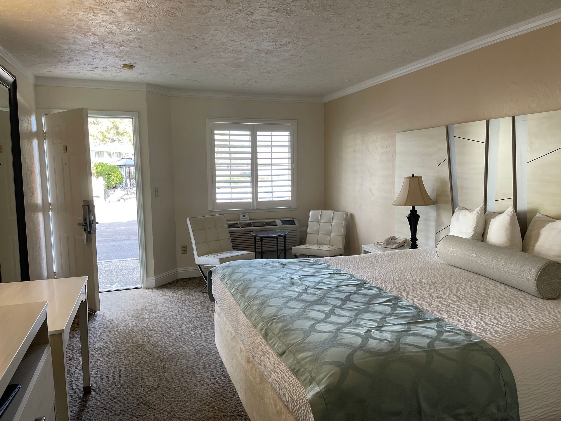 Hotel room with a bed, chairs, and an open door to a patio. Beige walls, carpet, and white shutters.