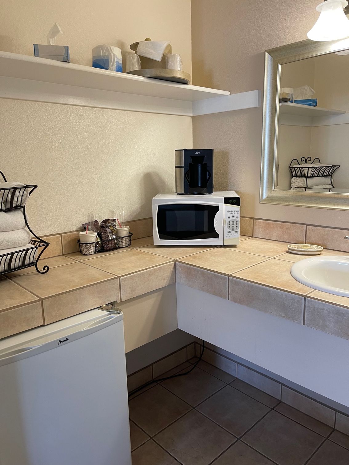 Bathroom counter with microwave, coffee maker, mini-fridge, and sink. Shelf with supplies above.