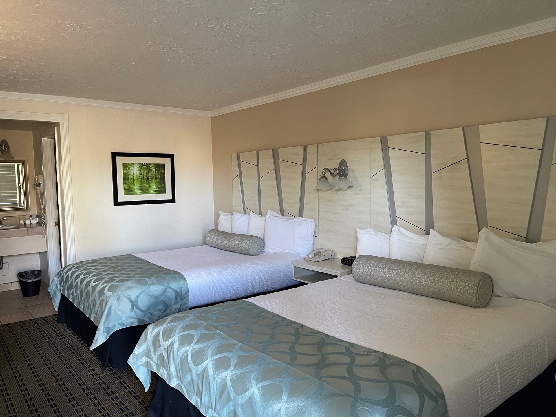 Two queen beds in a hotel room with teal bed runners, a framed picture, and a doorway to a bathroom.