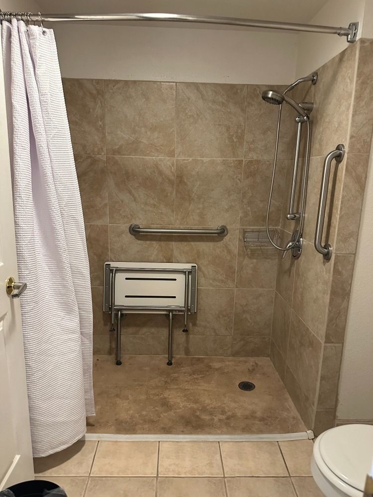 Accessible shower with a fold-down seat and grab bars. Cream-colored tiles, shower curtain, and toilet.