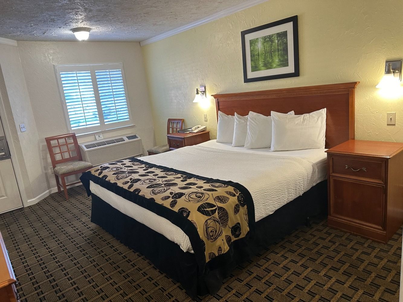 Hotel room with a bed, nightstand, window, and wall art. Dark wood furniture and neutral-colored walls.