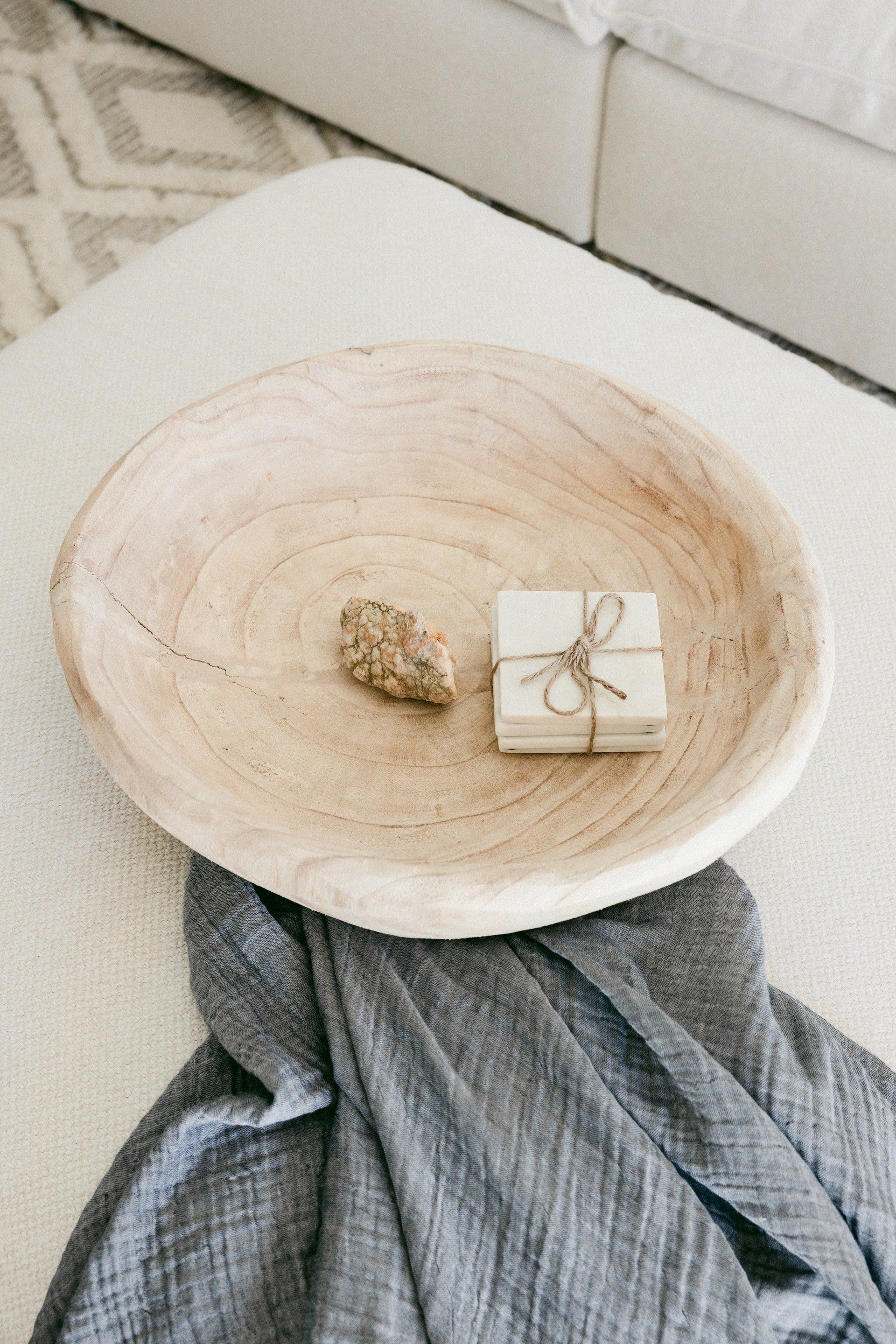 Wooden bowl with a small wrapped gift and a rock on a textured ottoman, next to a folded gray blanket.